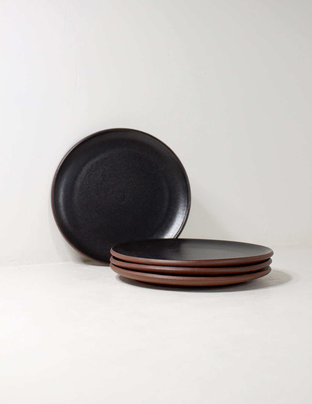 Hand-thrown stoneware coupe salad plates in matte black glaze, one upright behind a stack of three on white