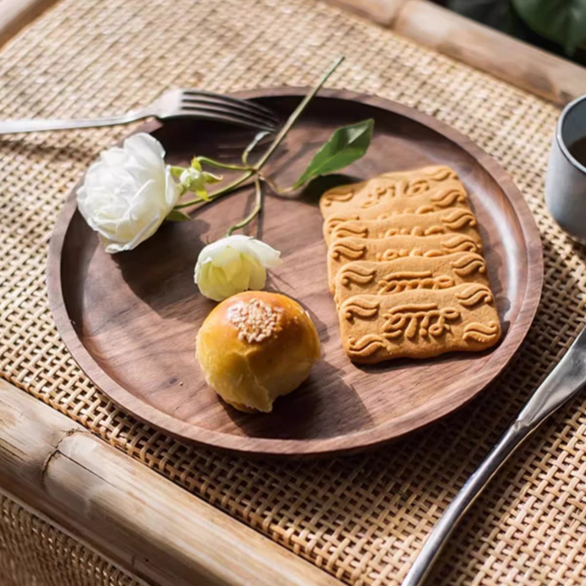 Round walnut serving tray set with a pastry, cookies and white roses on a woven rattan table runner