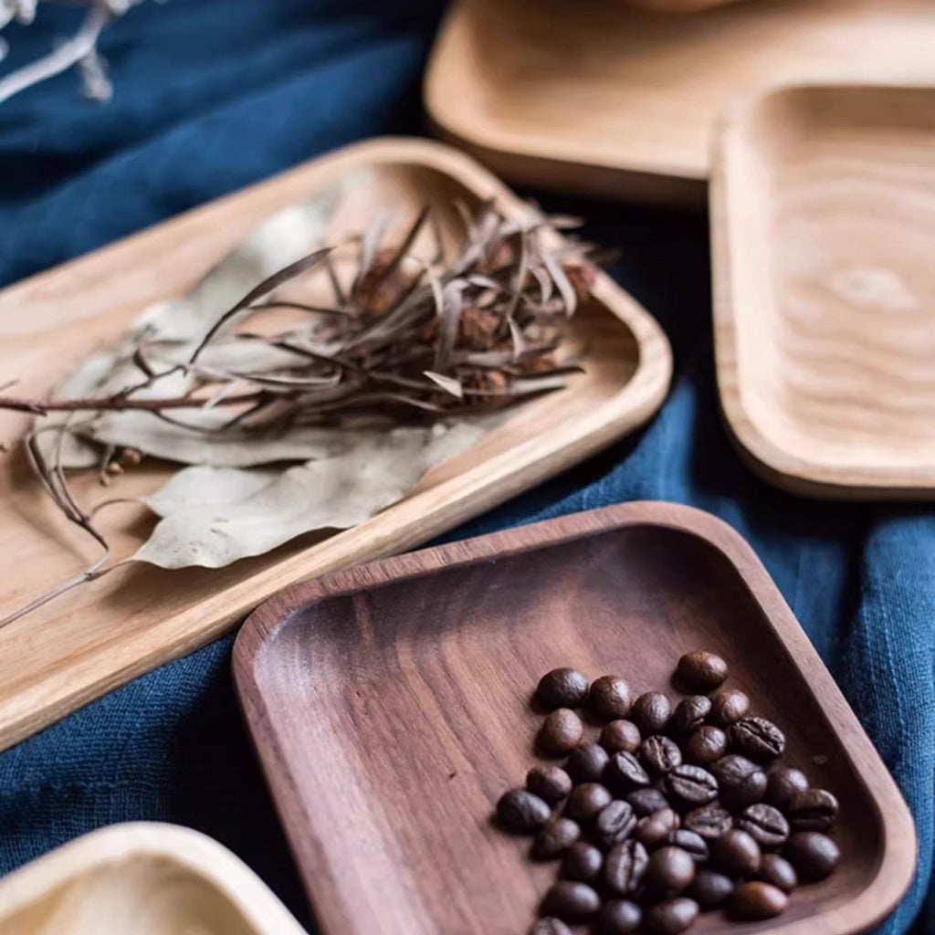 Small square walnut snack plate filled with coffee beans beside a long cherry plate of dried leaves