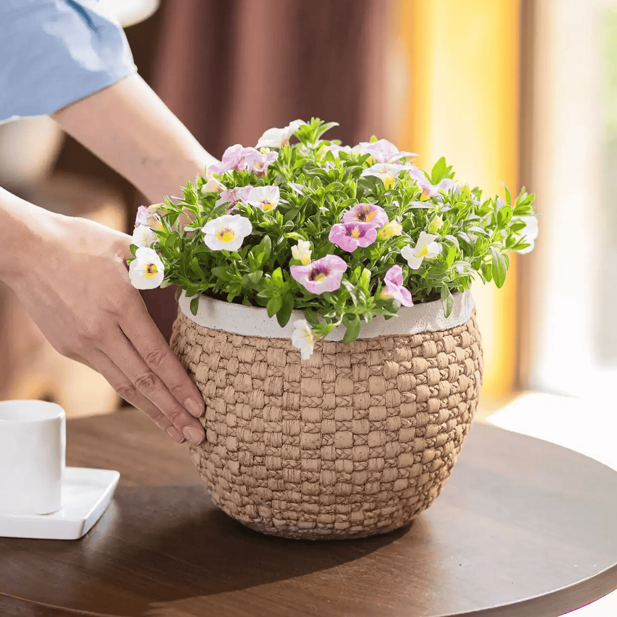 Hands placing a woven texture cement planter full of pink flowers on a round wooden table