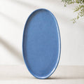 Large matte blue oval porcelain platter standing upright against a pale wall with a sprig of greenery