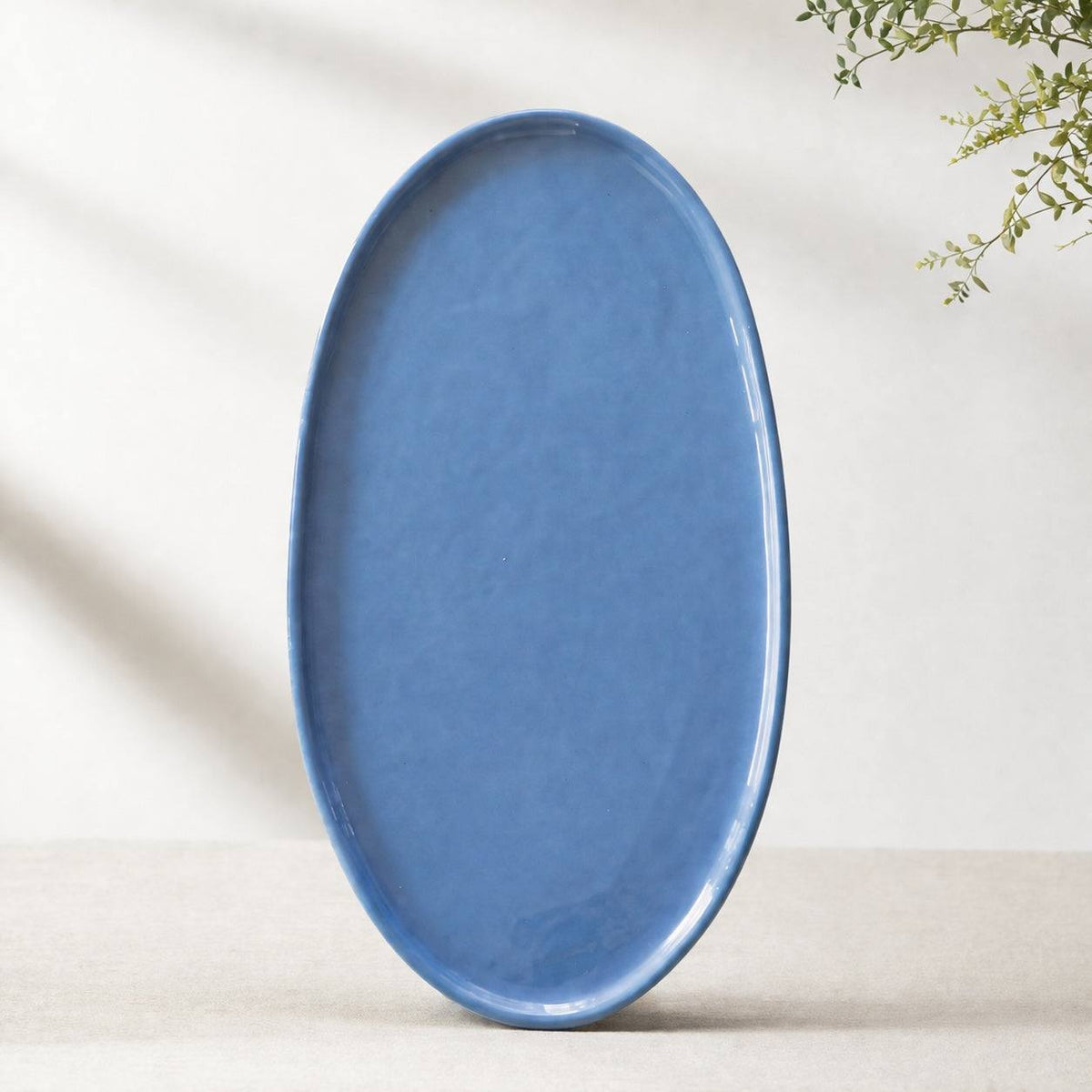 Large matte blue oval porcelain platter standing upright against a pale wall with a sprig of greenery