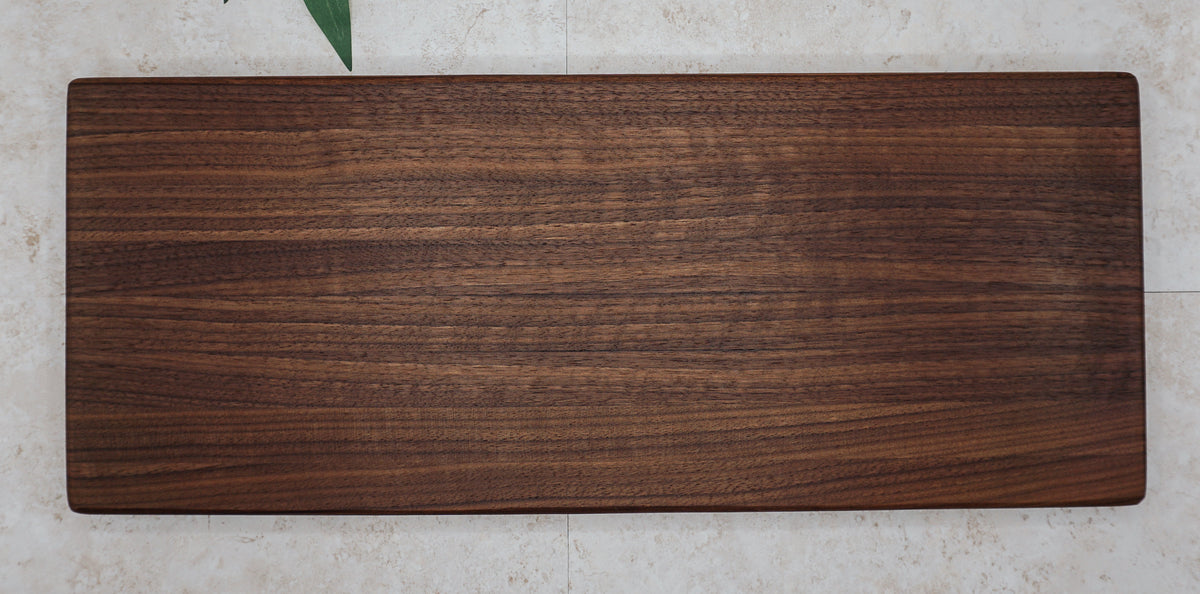Large dark walnut serving board seen from above on a pale stone surface with green leaves