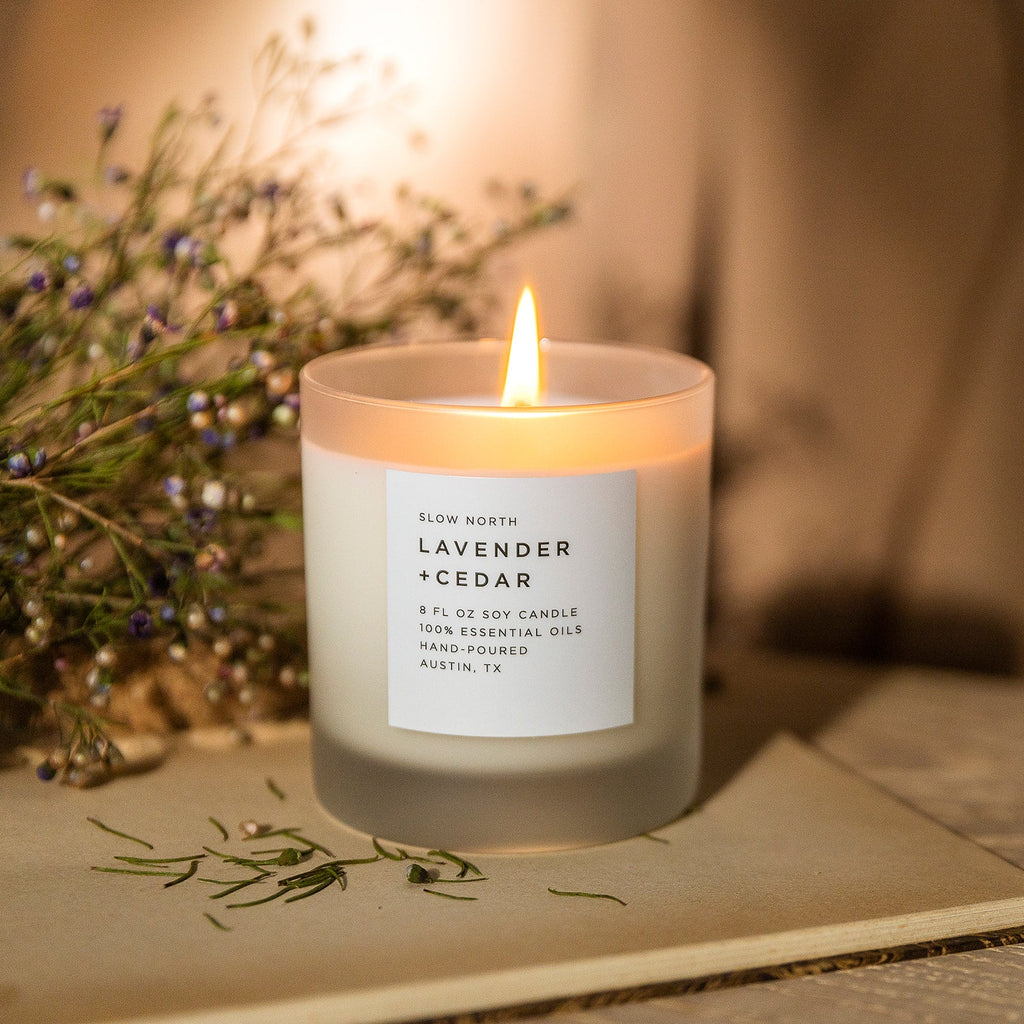 Lit lavender and cedar soy candle in frosted glass beside dried lavender stems on a linen-covered book