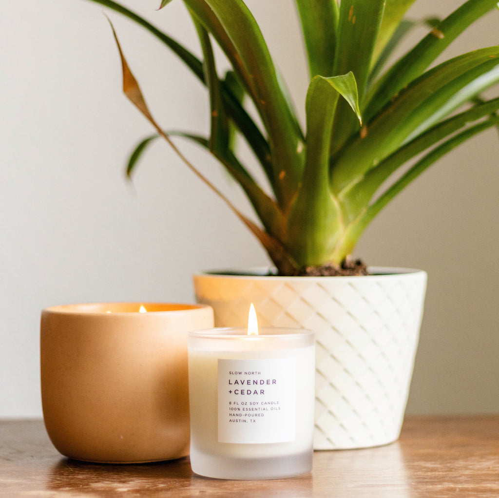 Lit frosted glass soy candle beside a tan candle and a potted plant in a white planter on a wood shelf