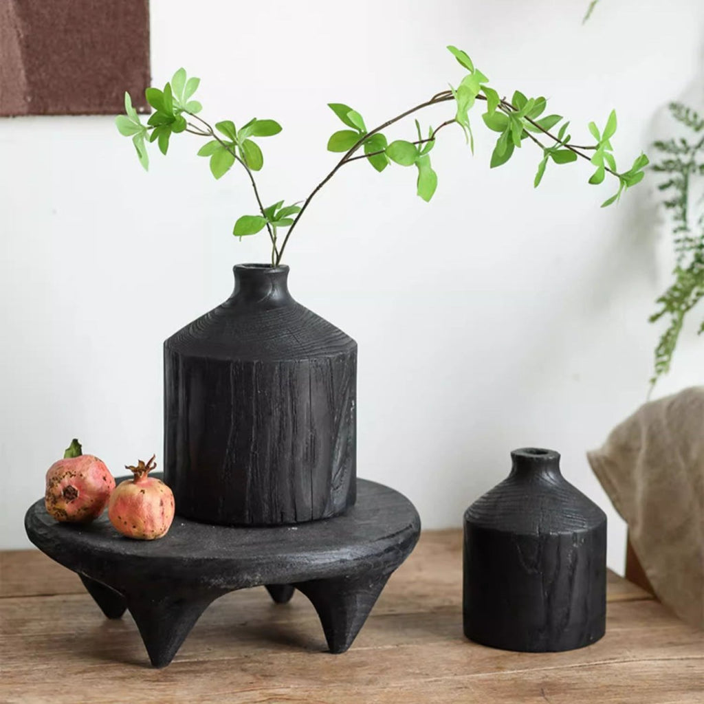 Medium black wood bottle vase with a green stem on a low black stand, small vase and pomegranates beside