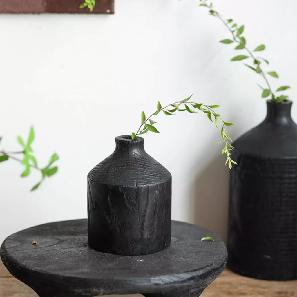 Small black wood bottle vase with a single trailing green stem on a round black wood stand