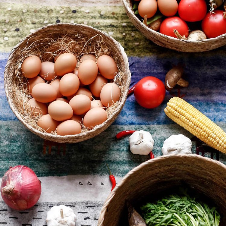 Round seagrass bowl basket of brown eggs on a striped rug, with tomatoes, corn and garlic around it