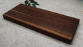 Dark edge-grain walnut serving board on a pale stone surface with green leaves at one corner