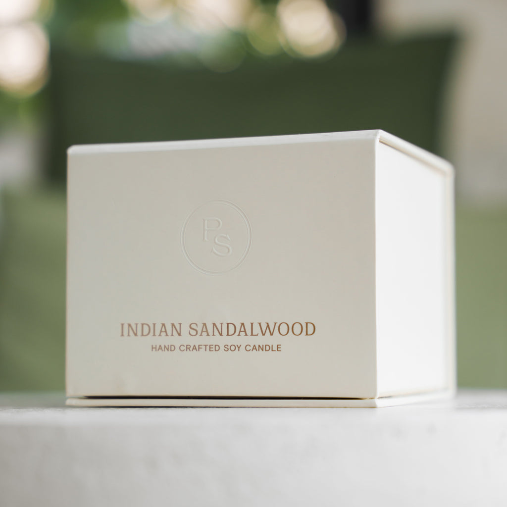 White Indian Sandalwood candle gift box with gold lettering on a pale ledge in front of green foliage