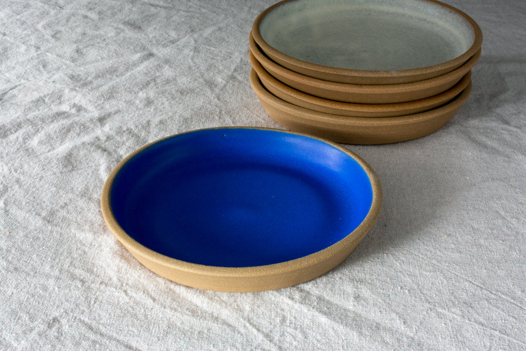 Fat Rim stoneware lunch plate in matte cobalt blue beside a stack of four oat-glazed plates on linen