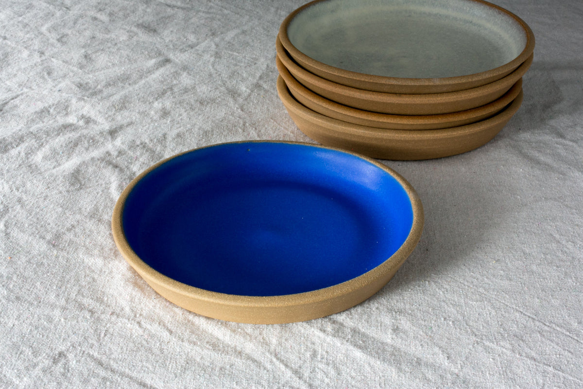 Fat Rim stoneware lunch plate in matte cobalt blue beside a stack of four oat-glazed plates on linen