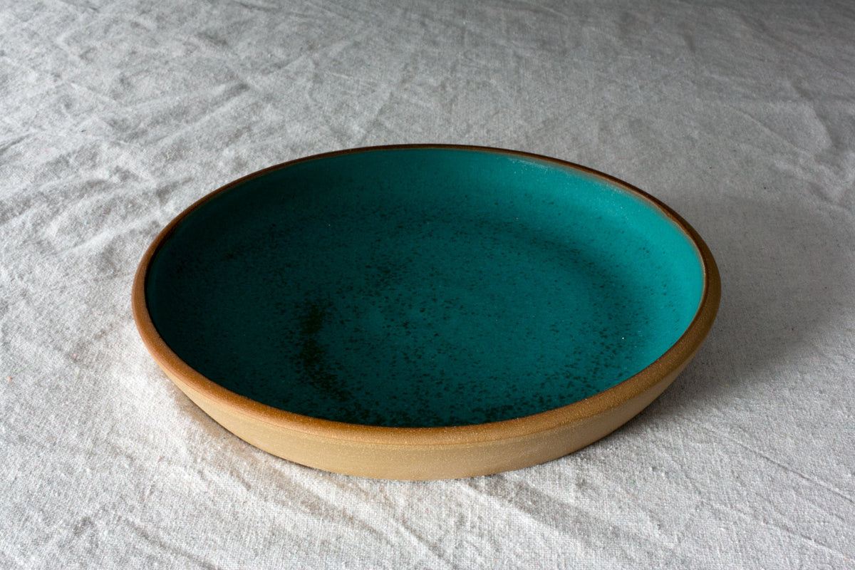 Fat Rim stoneware dinner plate in a deep speckled turquoise glaze with a clay rim on a white linen cloth