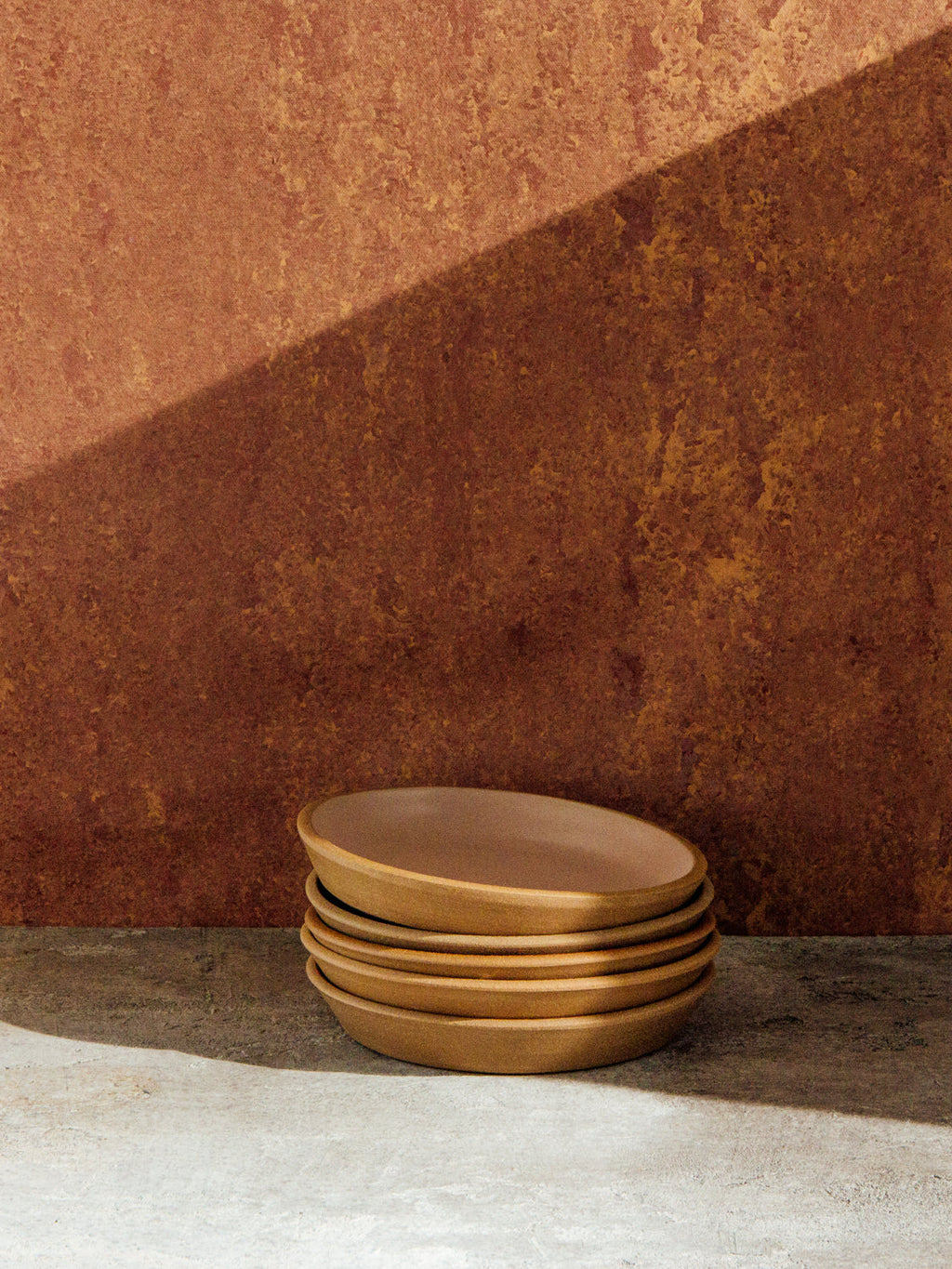 Stack of five Fat Rim stoneware plates in a sand glaze against a sunlit rust-colored plaster wall