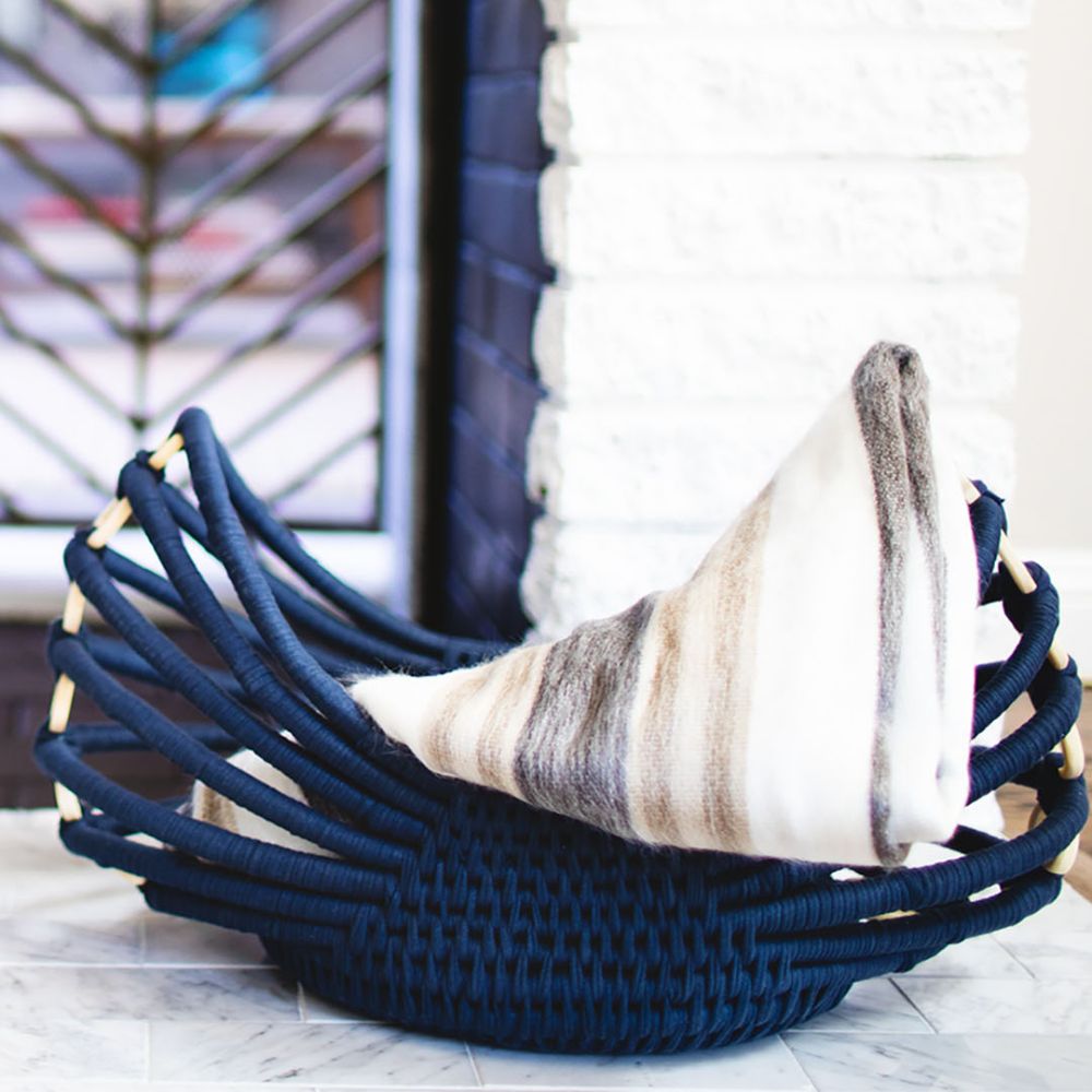 Folded striped alpaca throw resting in a dark blue woven basket by a white brick wall