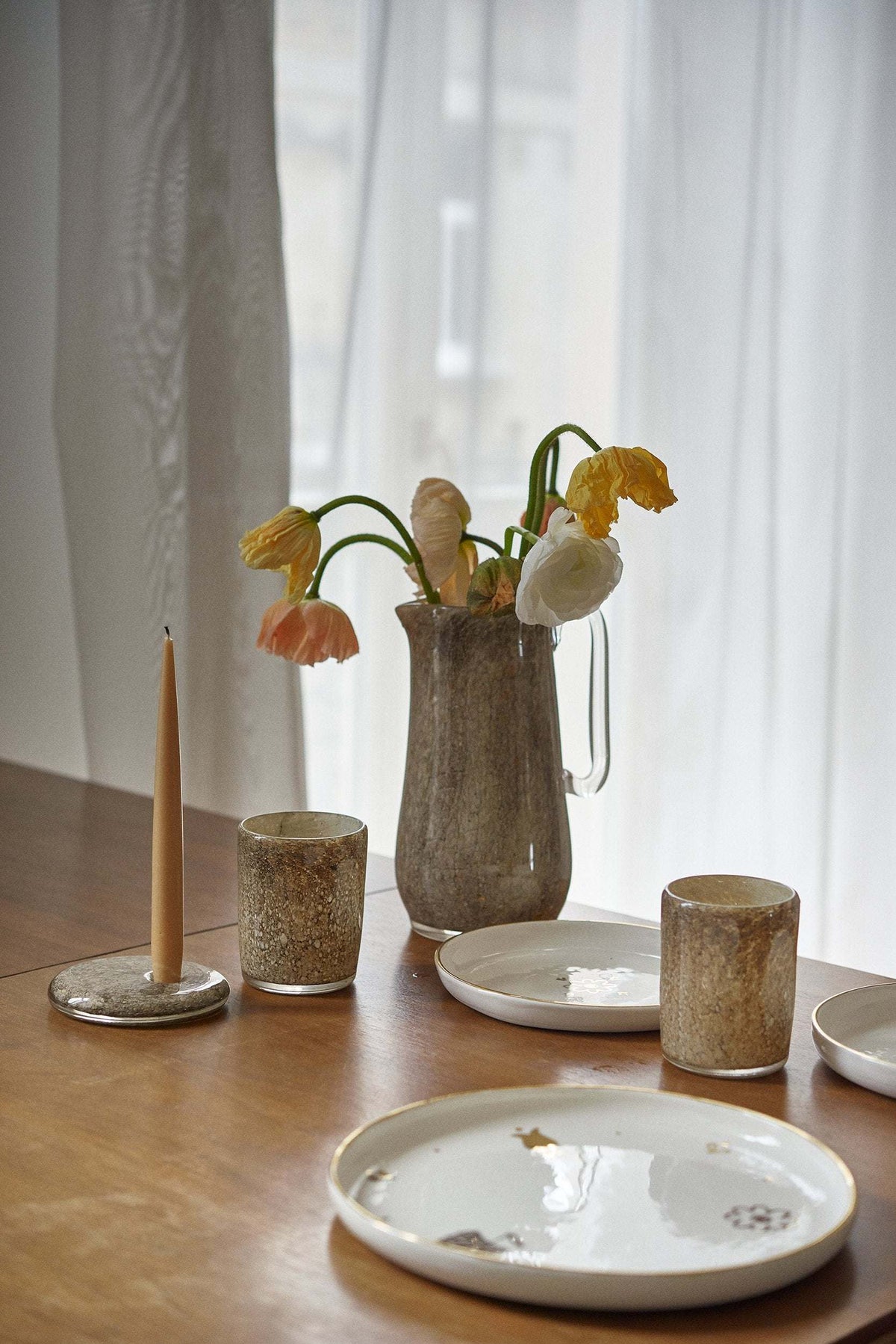 Marbled glass pitcher of spring flowers on a set wood table with candles and pale plates by a window