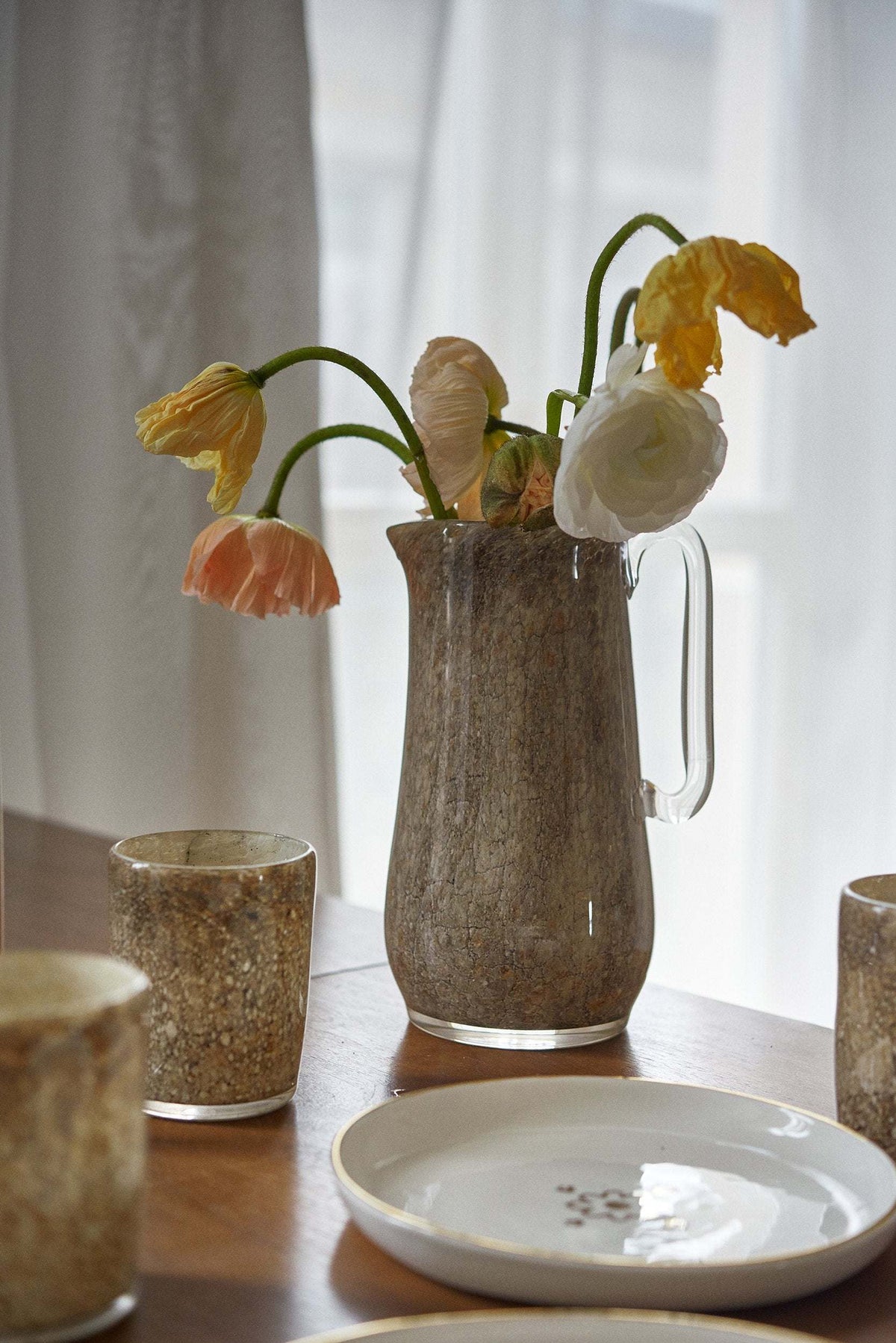 Hand-blown marbled glass pitcher with tulips beside white plates and amber glasses on a wood table