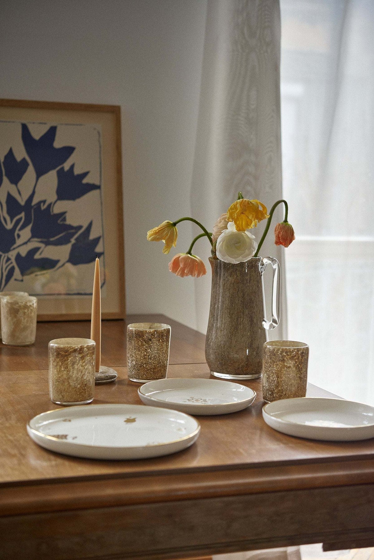 Wood table set with plates, glass cups and a marbled glass pitcher of tulips in front of blue artwork