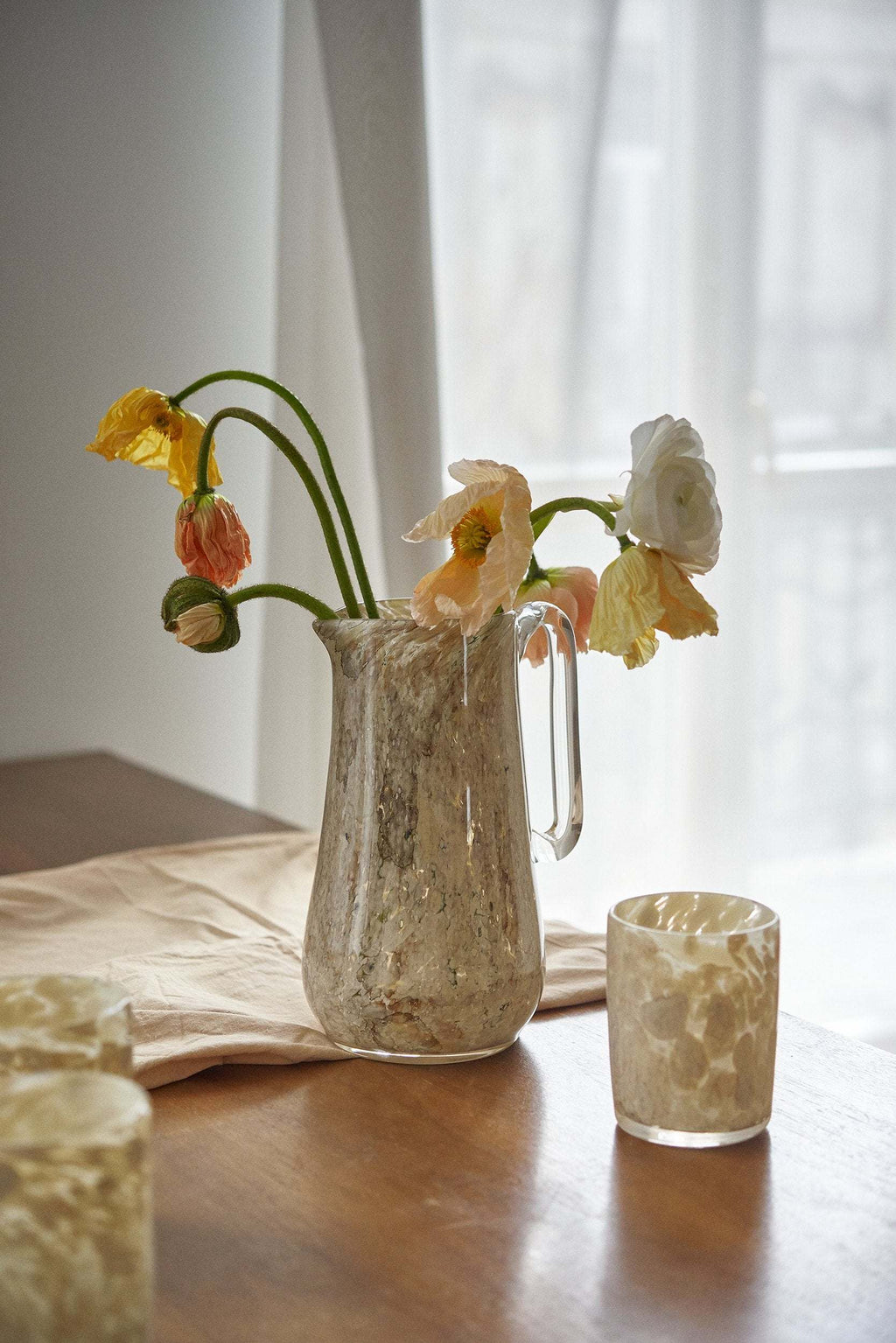 Marbled glass pitcher holding drooping yellow tulips on a wood dining table set with small glass cups