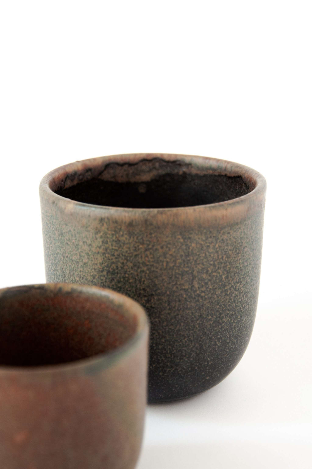 Close view of a handleless stoneware coffee cup with a rust glazed rim, a smaller rust cup blurred in front