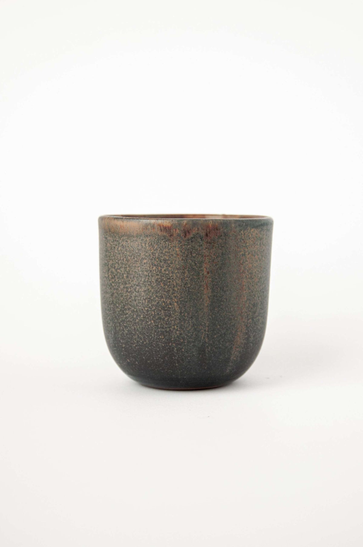 Handleless stoneware coffee cup in speckled grey green glaze with rust drips at the rim, side view on white