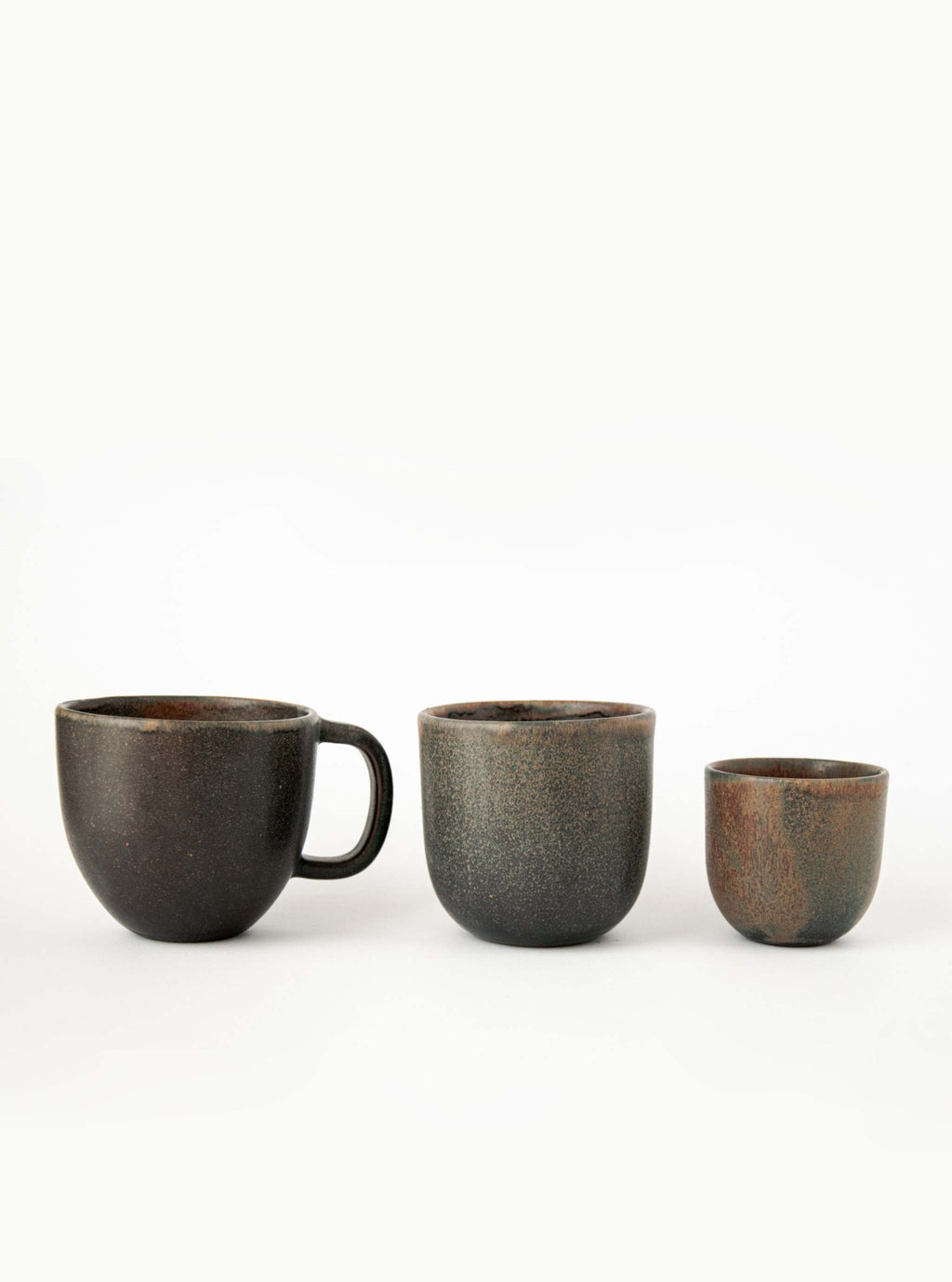 Handleless rust stoneware coffee cup in the center of a row between a handled mug and a small espresso cup