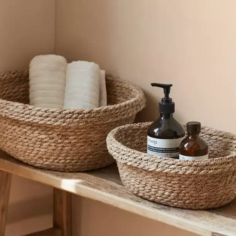 Open woven straw baskets with rolled towels and amber soap bottles on a sunlit wooden shelf