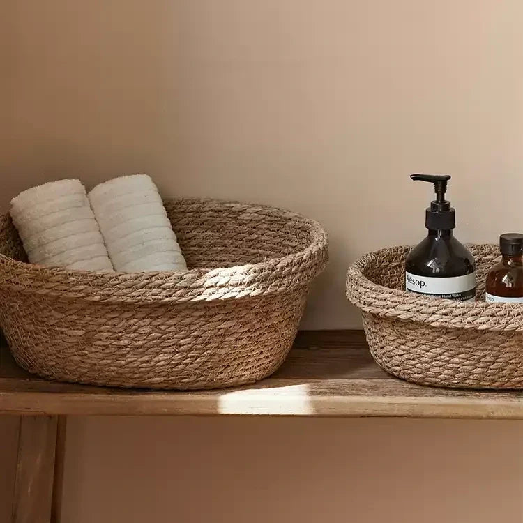 Open woven straw baskets in two sizes on a wooden shelf, one holding rolled white towels