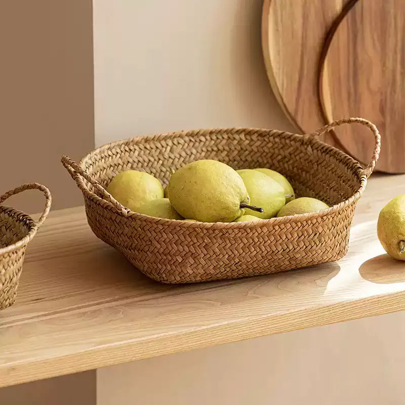 Oval seagrass bread basket holding pears on a pale wood shelf beside a round cutting board