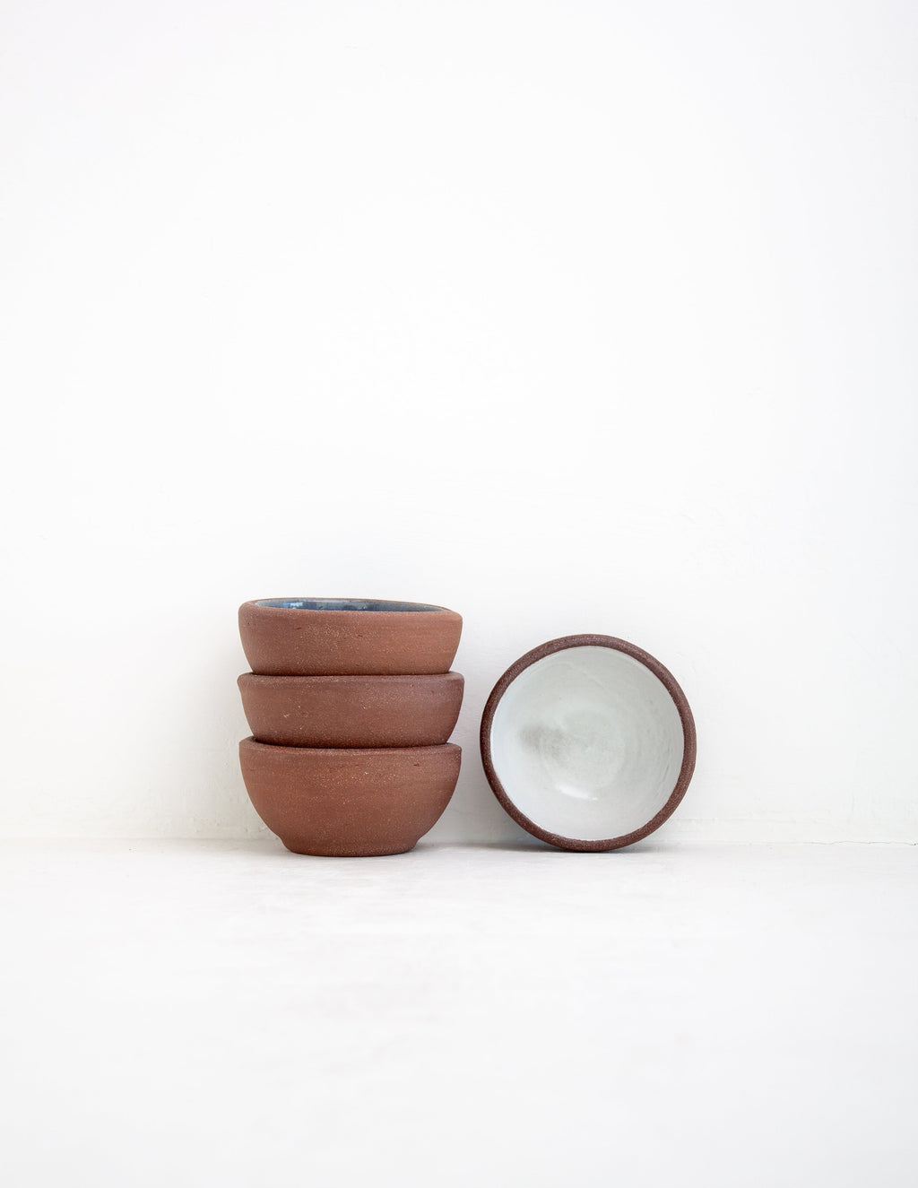 Stack of three hand-thrown stoneware sauce bowls with bare clay exteriors beside one tipped to show white glaze