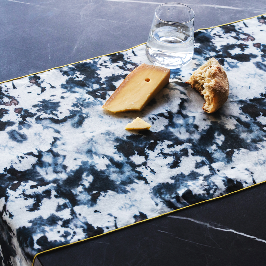 Grey marble print linen table runner with a water glass, cheese and bread on a black marble table