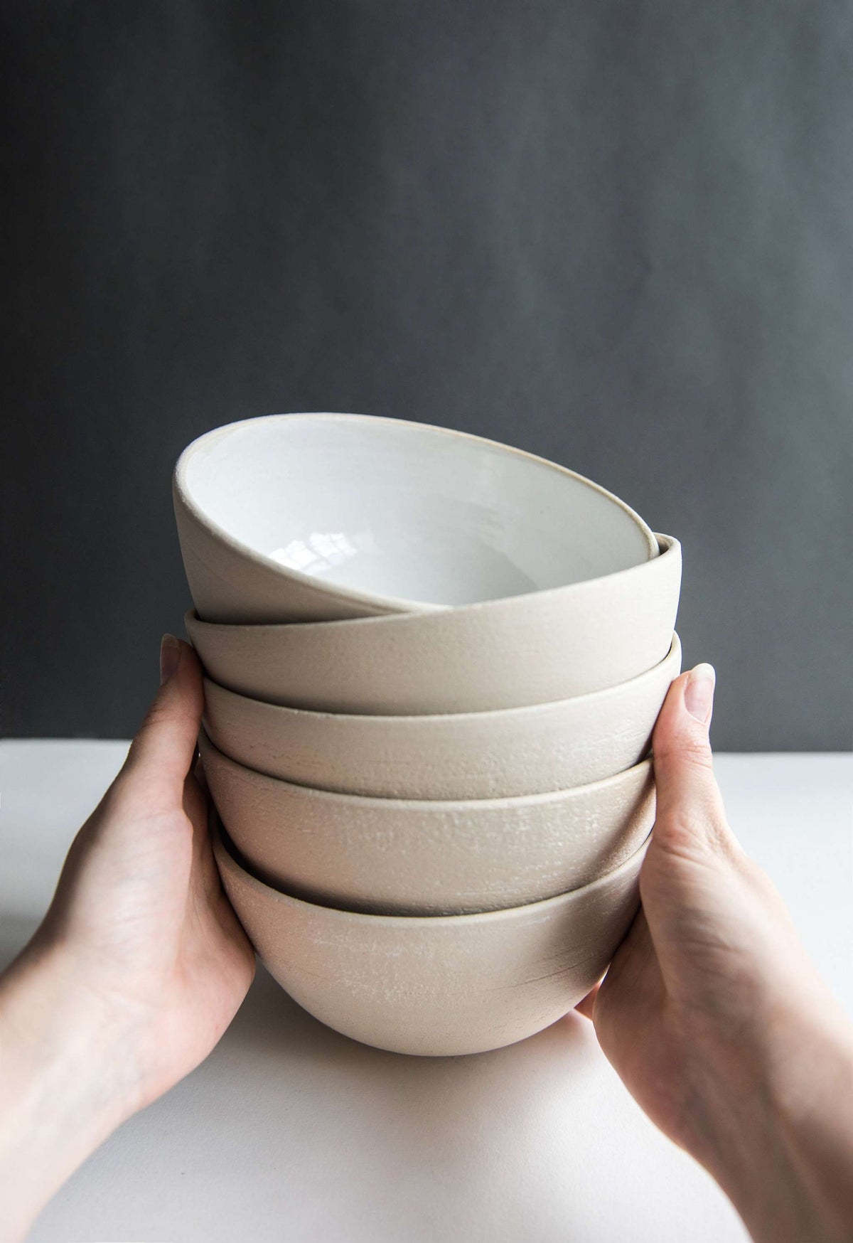 Two hands holding a stack of five exposed clay stoneware soup bowls with white glazed interiors for scale