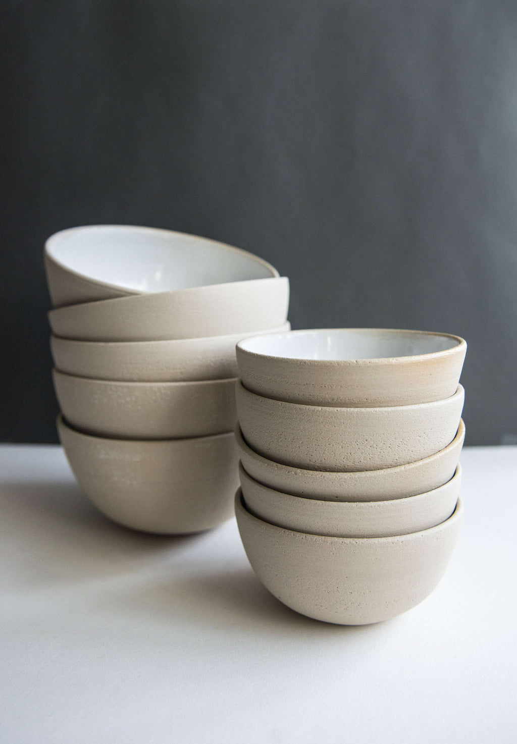 Two stacks of exposed clay stoneware bowls in two sizes side by side, white glazed interiors, charcoal backdrop