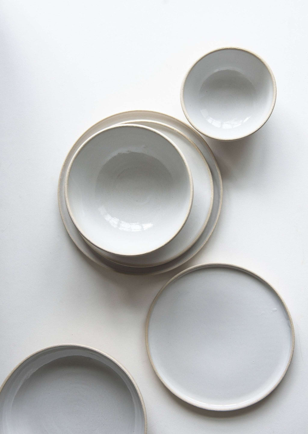 Overhead flat lay of white glazed stoneware bowls and plates with exposed clay rims arranged on a white surface