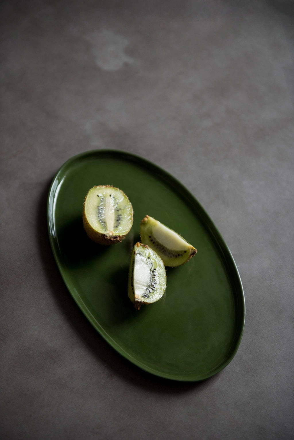 Handmade oval porcelain platter in dark green glaze holding three pieces of cut kiwi fruit on a grey stone surface