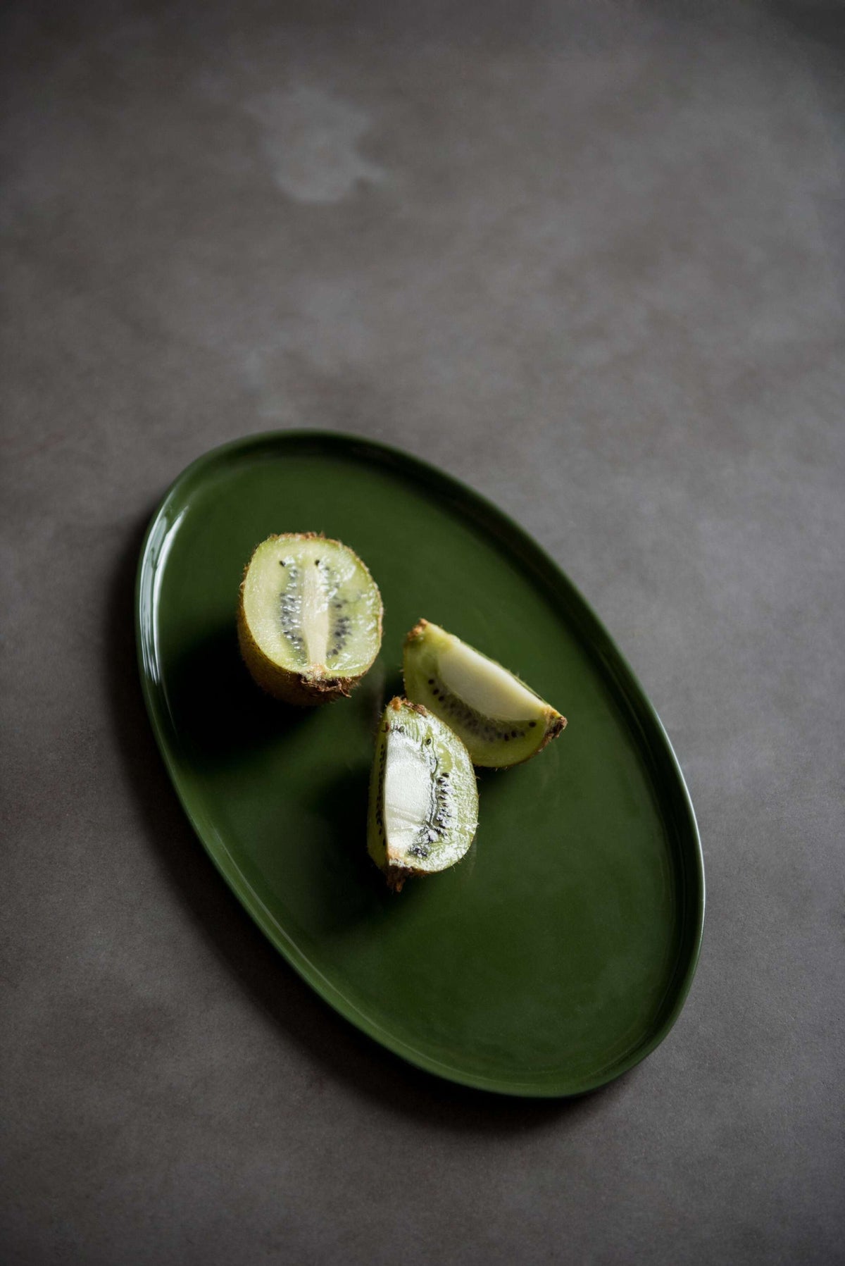 Handmade oval porcelain platter in dark green glaze holding three pieces of cut kiwi fruit on a grey stone surface