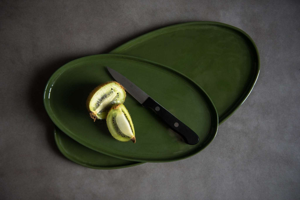 Two handmade oval porcelain platters in green glaze stacked crosswise with sliced kiwi and a black handled knife