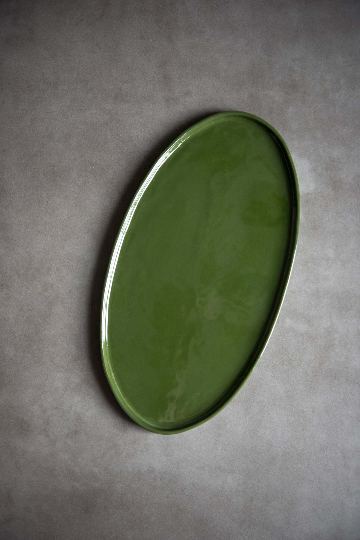 Handmade oval porcelain platter in green glaze set at an angle, light catching the raised rim, on a grey surface