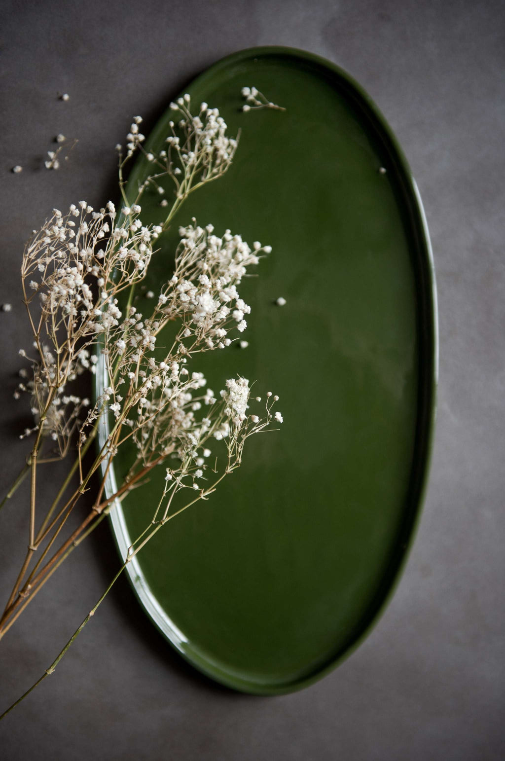 Handmade oval porcelain platter in glossy deep green glaze with dried baby's breath stems laid across it, overhead
