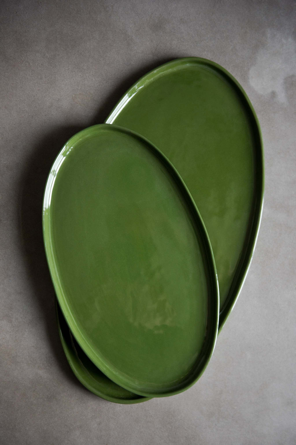 Two handmade oval porcelain platters in glossy green glaze, overlapping in two sizes on a grey stone surface