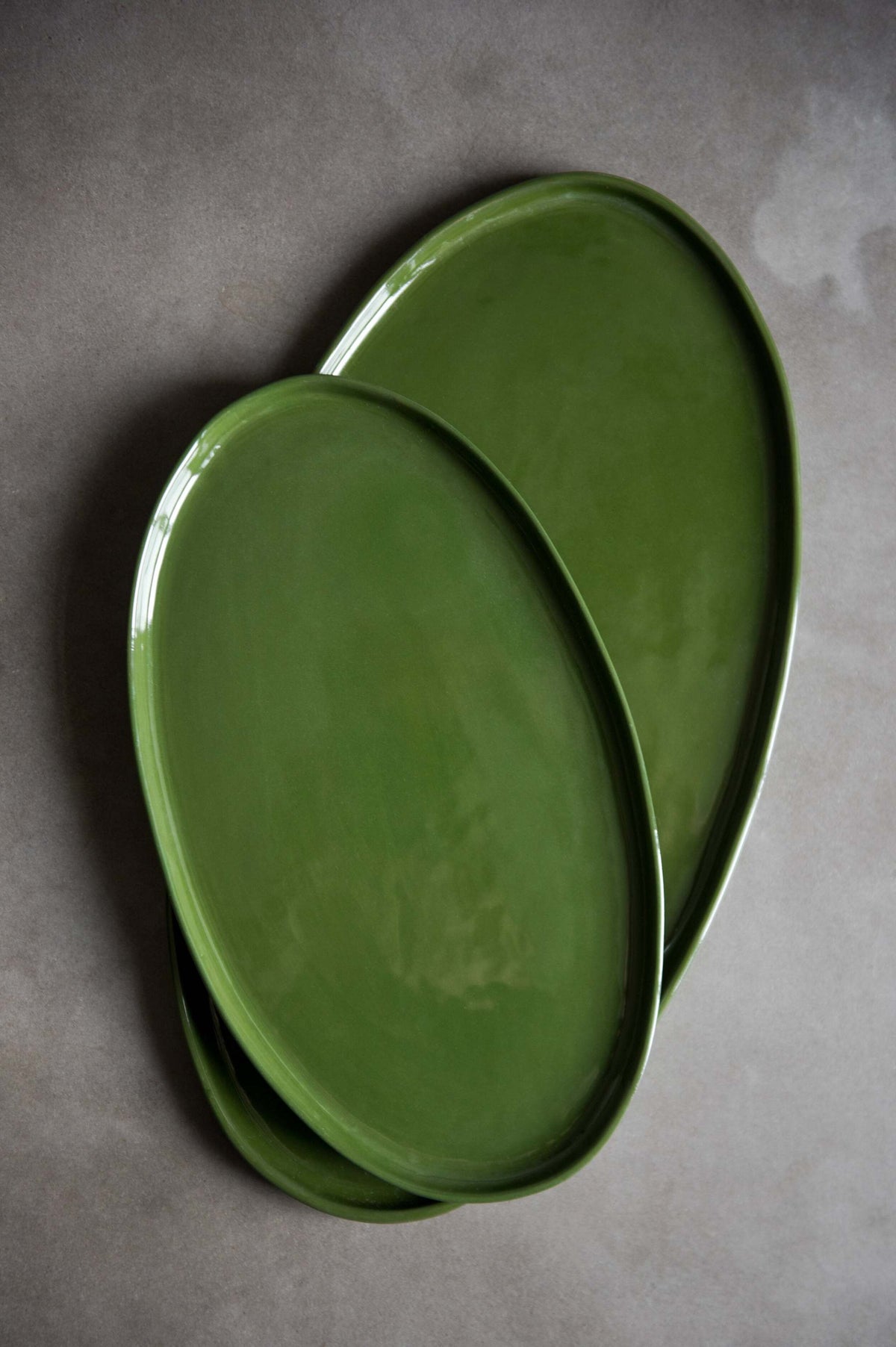 Two handmade oval porcelain platters in glossy green glaze, overlapping in two sizes on a grey stone surface