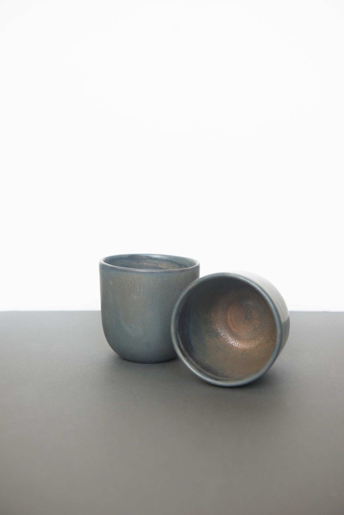 Two handleless stoneware coffee cups in blue grey glaze, one tipped on its side to show the rust brown interior