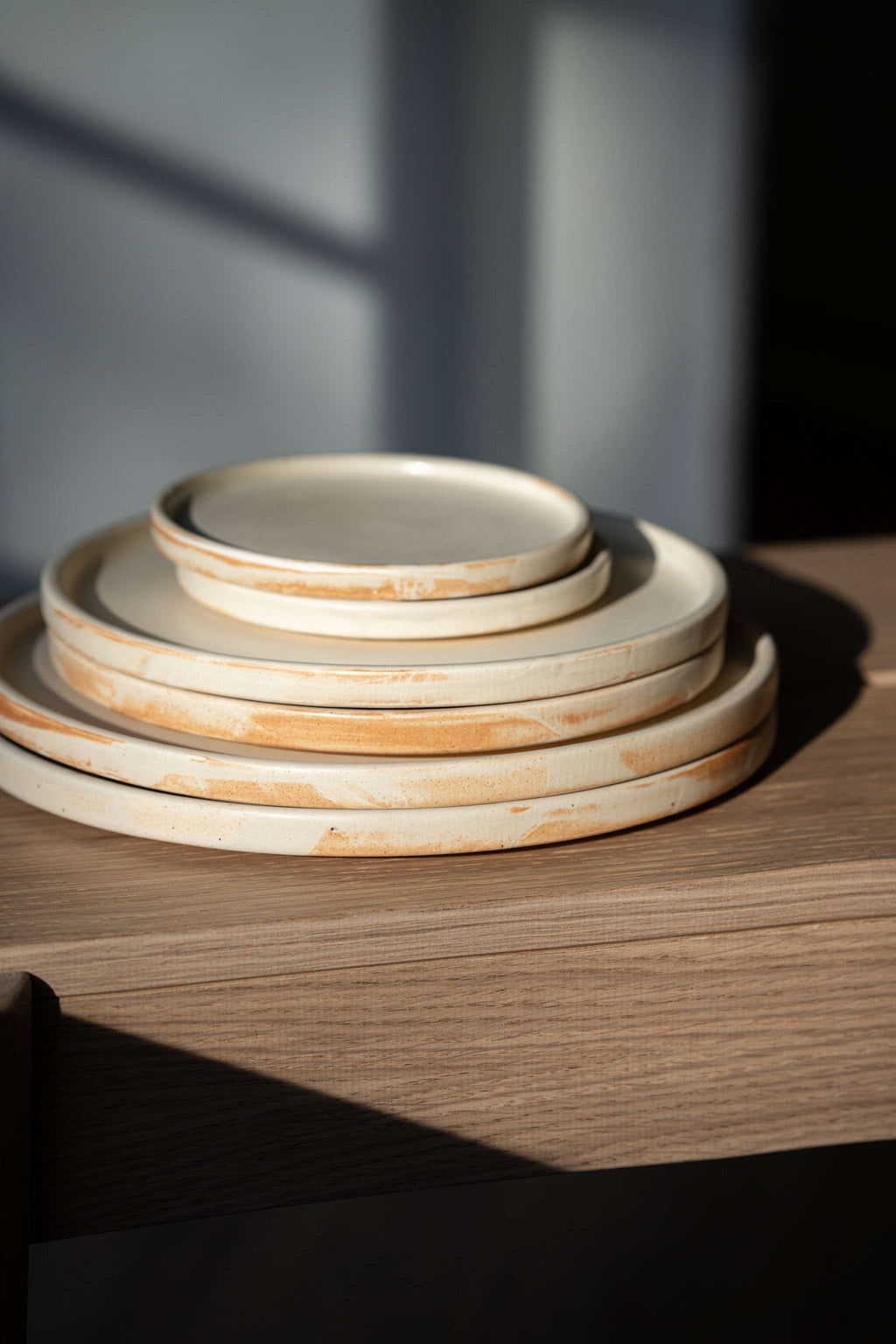 Six beige matte stoneware plates stacked by size on an oak bench, side view showing rust brushed edges