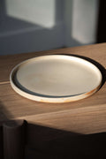 Single beige matte stoneware dinner plate with a low straight rim on an oak bench in late afternoon light