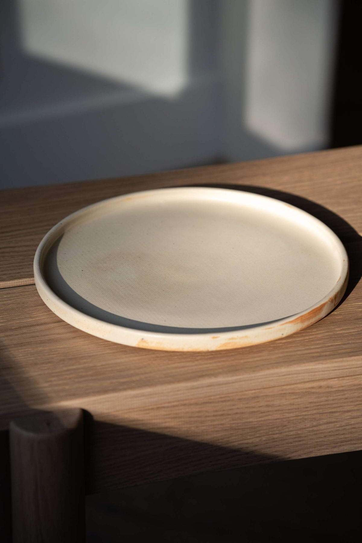Single beige matte stoneware dinner plate with a low straight rim on an oak bench in late afternoon light