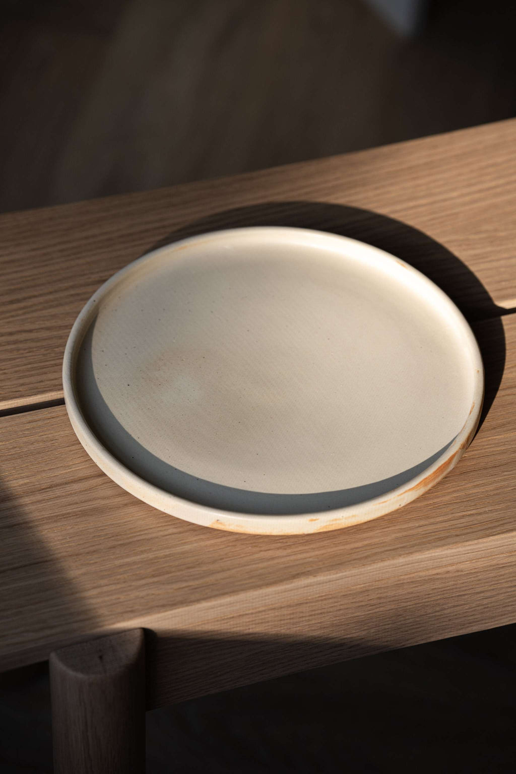 Beige matte stoneware dinner plate seen from a high angle on a slatted oak bench, casting a long shadow