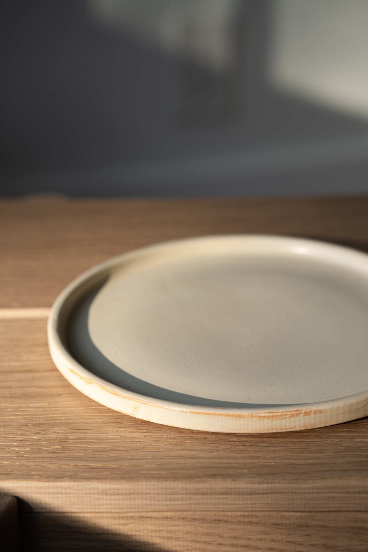 Close-up of the raised rim on a beige matte stoneware dinner plate with faint rust streaks, on an oak bench