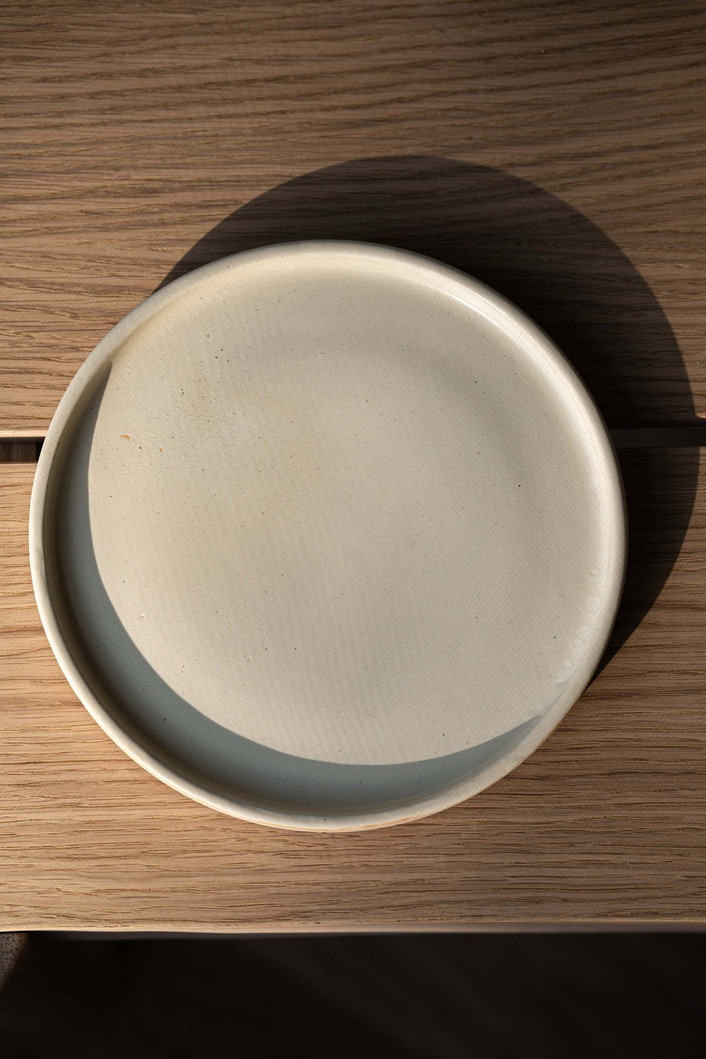 Beige matte stoneware dinner plate from directly above on oak, a curved shadow falling across its surface