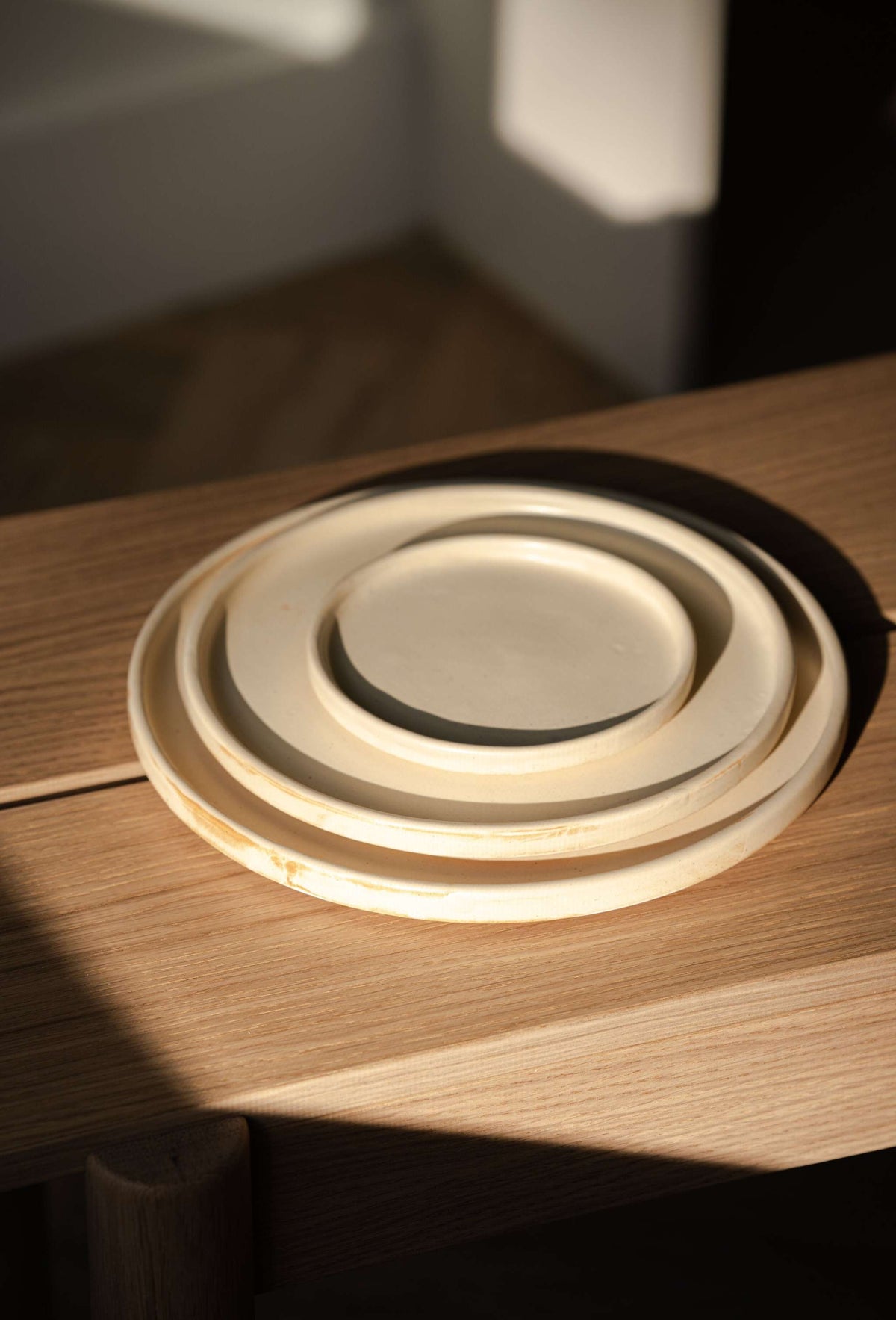 Three beige matte stoneware plates nested by size on an oak bench, angled view in soft window light