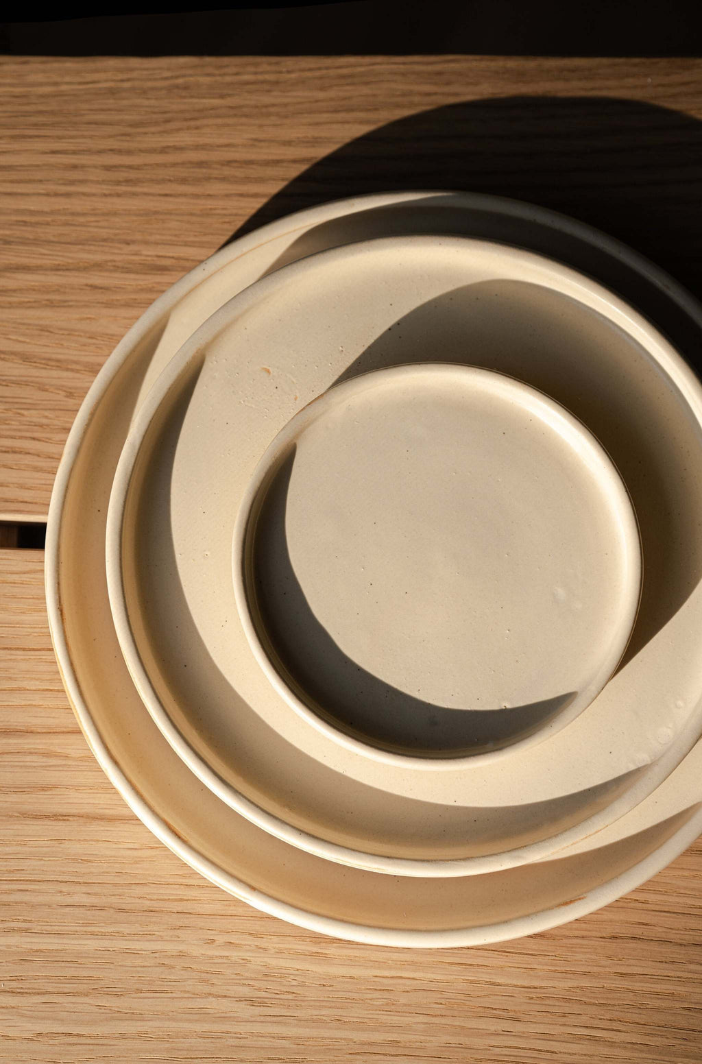Overhead close view of three nested beige matte stoneware plates with shadows curving inside each rim